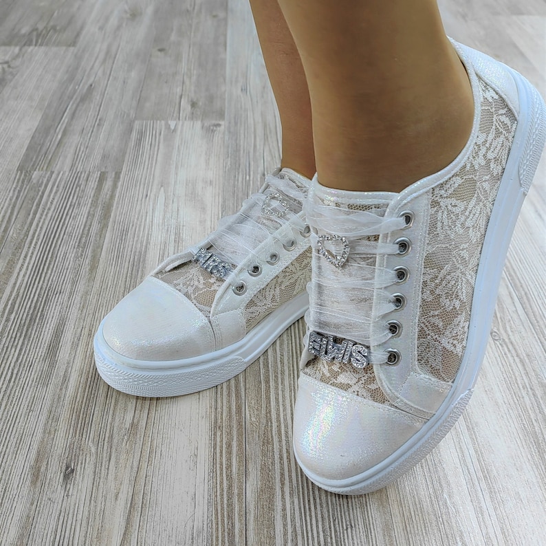 K&ouml;nnte beinhalten: Ein Paar wei&szlig;e Sneaker mit floralem Spitzen&uuml;berzug an den Seiten, schillernden Zehenkappen und transparenten Schleifenb&auml;ndern. Jeder Schuh ist mit einem silbernen herzf&ouml;rmigen Anh&auml;nger und einem silbernen Strass-Anh&auml;nger mit dem Wort ELVIS verziert.