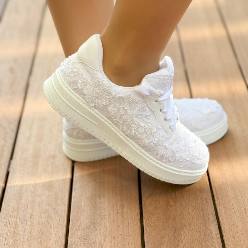 May include: White lace sneakers with a white satin ribbon and a white lace design. The sneakers are on a wooden surface.