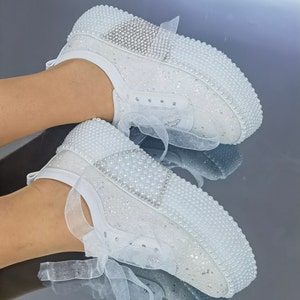 May include: White sneakers with pearl embellishments and a white tulle ribbon. The sneakers have a sparkly finish.