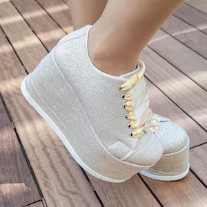 Könnte beinhalten: Ein Paar silberne Glitzer-Plateau-Sneaker mit einer dicken weißen Sohle und breiten cremefarbenen Satinband-Schnürsenkeln. Die Schuhe sind mit einem kleinen, kreisförmigen, mit Kristallen besetzten dekorativen Akzent am oberen Schnürbereich verziert.