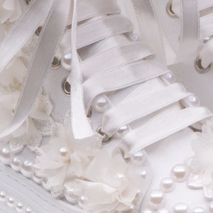 May include: White canvas high-top sneakers with white satin ribbon laces and pearl embellishments. The sneakers are decorated with white fabric flowers and pearls.
