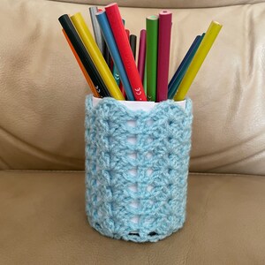 Peut inclure: Un pot à crayons bleu clair en crochet rempli de crayons de couleur. Le support présente un motif ajouré décoratif et repose sur une surface beige. Les crayons sont disposés verticalement, prêts à l'emploi.