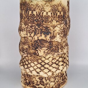 Op de afbeelding: Een beige keramische vaas met ingewikkelde, donkerbruine geëtste ontwerpen. De vaas heeft gedetailleerde patronen van bloemen, geometrische en sierlijke motieven. De bovenrand heeft een geschulpte rand en het algehele ontwerp is getextureerd.