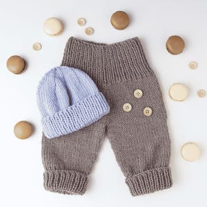 Puede incluir: Un pantalón de punto marrón con cintura alta acanalada y dobladillos, con tres pequeños botones de madera en la parte delantera, acompañado de un gorro de punto azul claro, rodeado de varias galletas redondas pequeñas y botones.