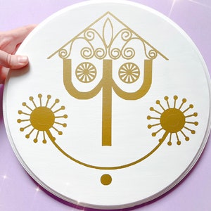 May include: A round, white wooden sign featuring a gold-colored stylized face design. The design includes a house-shaped top with swirls, two sun-like shapes, and a smiling mouth. The sign is held by a hand.