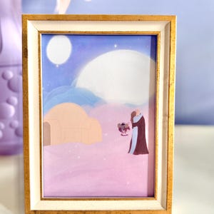 May include: A rectangular picture frame with a gold and white border containing an illustration of two figures embracing in a desert landscape under a large moon and a smaller star, with a domed structure nearby on a pink and purple ground.