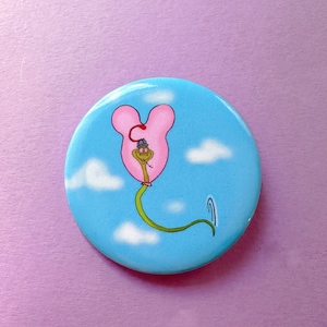 Pode incluir: Um botão azul e rosa com uma ilustração de desenho animado de uma criatura verde usando um chapéu e andando dentro de um balão rosa em forma de coração. O balão flutua em um céu azul com nuvens brancas.