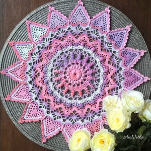 May include: A handmade, star-shaped crochet doily in shades of pink, purple, and white. The intricate design features a central spiral, surrounded by layers of detailed patterns and heart-shaped points. The doily rests on a gray placemat, with yellow roses in the foreground.