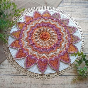 May include: A handmade, colorful crochet doily. The doily features a circular design with intricate patterns in shades of orange, pink, and beige. The outer edge has a scalloped trim. The doily is placed on a white placemat with a gold trim.