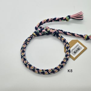 May include: A blue, pink, and green braided bracelet with a brown tag that says "KUMIHIMO 組み紐". The bracelet is tied in a knot and has a loose end on each side. The tag is labeled "K8".