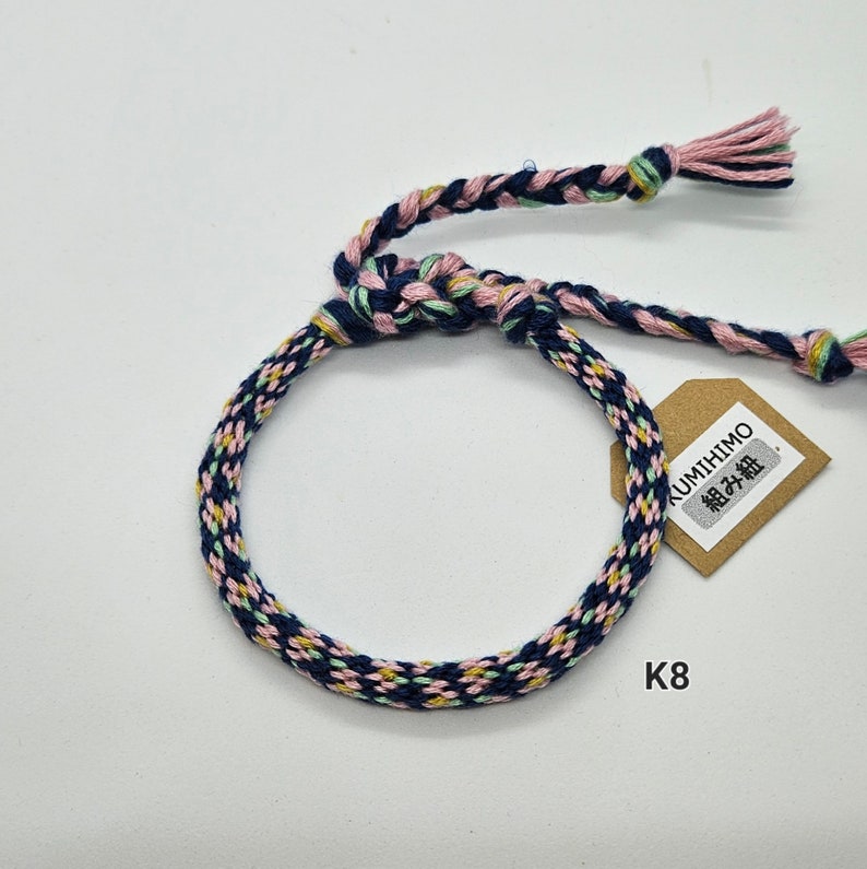 May include: A blue, pink, and green braided bracelet with a brown tag that says "KUMIHIMO 組み紐". The bracelet is tied in a knot and has a loose end on each side. The tag is labeled "K8".