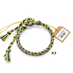 May include: A yellow, black, and turquoise braided bracelet with a brown tag that says "KUMIHIMO 組紐" and "K3".