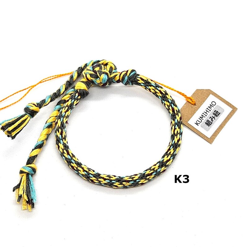 May include: A yellow, black, and turquoise braided bracelet with a brown tag that says "KUMIHIMO 組紐" and "K3".