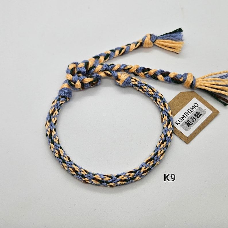 May include: A braided bracelet with blue, yellow, and green threads. The bracelet is tied with a knot and has a small tag that says "KUMITHIMO 組紐".