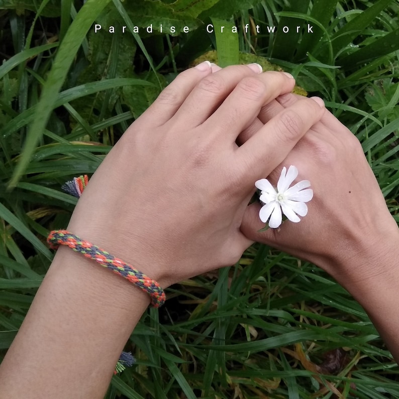 May include: A close-up of two hands holding a white flower. The hands are wearing a colorful braided bracelet with a green, orange, and gray pattern. The text "Paradise Craftwork" is visible in the background.