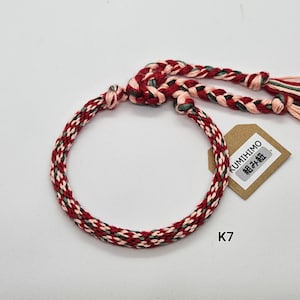 May include: Red, pink, and green braided bracelet with a KUMIHIMO tag that says "組み紐". The tag is attached to the bracelet with a knot. The bracelet is labeled K7.