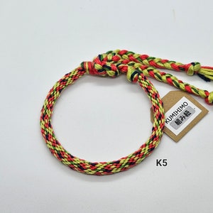 May include: A multi-colored braided bracelet with red, yellow, green, and blue threads. The bracelet is tied with a knot and has a small tag that says "KUMIHIMO 組み紐".