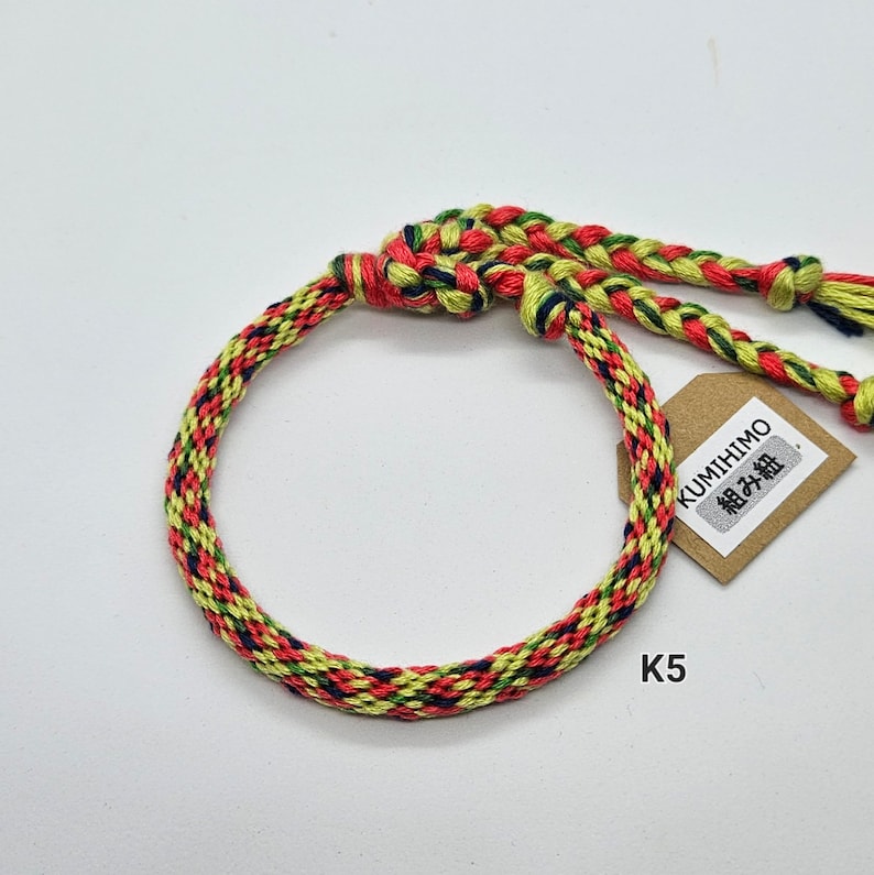 May include: A multi-colored braided bracelet with red, yellow, green, and blue threads. The bracelet is tied with a knot and has a small tag that says "KUMIHIMO 組み紐".