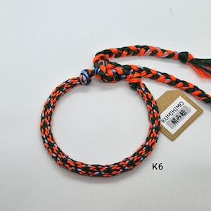 May include: A braided bracelet in orange, green, and blue colors. The bracelet is made with a technique called "Kumihimo" and has a tag that says "Kumihimo" in Japanese characters. The tag also has the text "組み紐" in Japanese characters. The bracelet is labeled "K6".