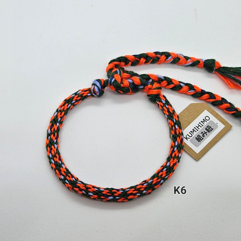 May include: A braided bracelet in orange, green, and blue colors. The bracelet is made with a technique called "Kumihimo" and has a tag that says "Kumihimo" in Japanese characters. The tag also has the text "組み紐" in Japanese characters. The bracelet is labeled "K6".