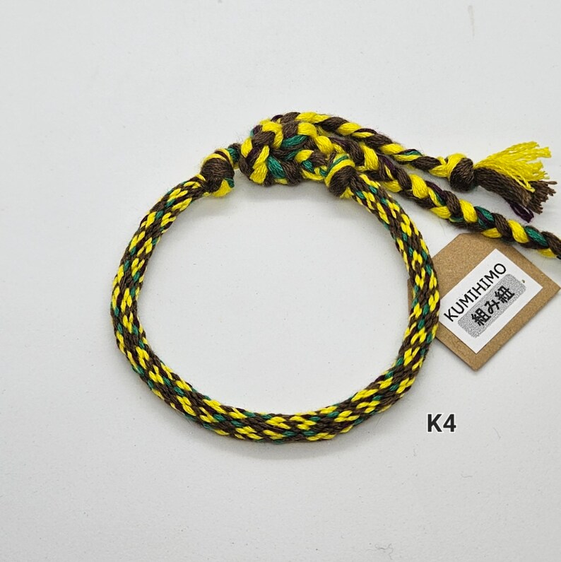 May include: A yellow, green, and brown braided bracelet with a brown tag that says "KUMIHIMO 組み紐".