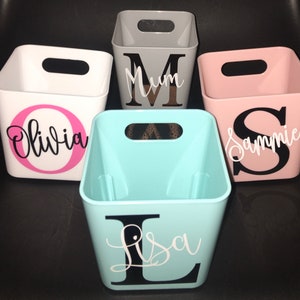 May include: Four square storage bins with handles. The bins are white, grey, pink, and light blue. Each bin has a letter and name on the front. The names are Olivia, Mum, Sammie, and Lisa.