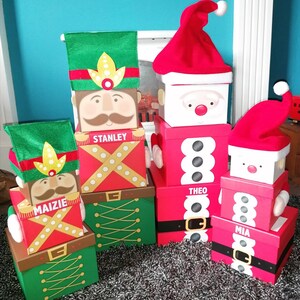 May include: Four red and green Christmas gift boxes with Santa and toy soldier designs. The boxes are stacked and labelled with the names "Mia", "Theo", "Stanley", and "Maizie".