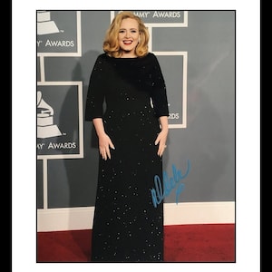 Puede incluir: Un vestido negro de noche largo con una textura brillante. La mujer que lleva el vestido tiene el pelo rubio y sonríe. El fondo es una pared gris con las palabras "GRAMMY AWARDS" impresas en ella. La mujer ha firmado su nombre en tinta azul en la imagen.