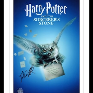 Puede incluir: Un póster enmarcado de "Harry Potter y la piedra filosofal" con un fondo azul. Un búho lleva una carta, con el título en la parte superior y el logotipo de Wizarding World en la parte inferior. Incluye una firma.