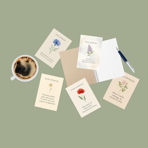 Puede incluir: Una vista cenital de un espacio de trabajo sereno con tarjetas con ilustraciones florales y texto en polaco, una taza de café, un cuaderno y un bolígrafo. Las tarjetas muestran varias flores como un aciano azul, una margarita, una amapola roja y una rosa rosa.