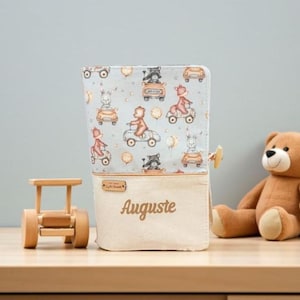 Peut inclure: Une trousse rectangulaire en tissu sur fond bleu clair avec un motif d'animaux illustrés conduisant des voitures et des scooters. La partie inférieure est en toile beige avec le nom Auguste brodé en lettres cursives marron.
