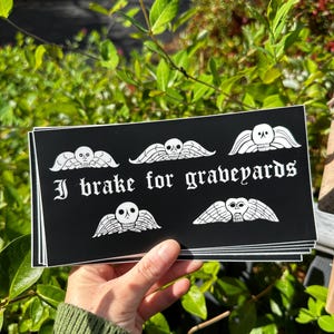 May include: A set of five black and white stickers with a skull and wings design. The stickers say "I brake for graveyards".