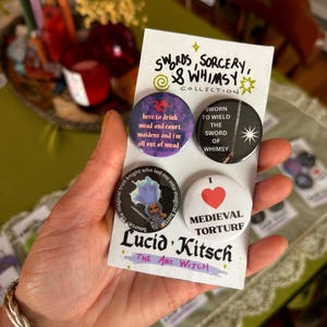 May include: A set of four circular pin-back buttons on a white display card labeled Swords, Sorcery, & Whimsy Collection and Lucid Kitsch The Art Witch. The buttons feature various text and illustrations, including a troll knight and a red heart.