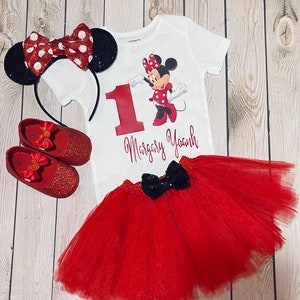 May include: A red and white Minnie Mouse themed first birthday outfit for a baby girl. The outfit includes a white bodysuit with a red number one, a red tulle tutu skirt, a red headband with black ears and a red bow, and red glitter shoes.