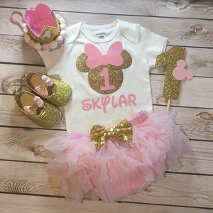 Puede incluir: Un conjunto de primer cumplea&ntilde;os rosa y dorado para una ni&ntilde;a. El conjunto incluye un body blanco con un n&uacute;mero uno de purpurina dorada y el nombre "SKYLAR" en purpurina rosa, una falda de tul rosa con un lazo de purpurina dorada y zapatos de purpurina dorada con lazos rosas. El conjunto tambi&eacute;n incluye una corona de purpurina rosa y dorada y un n&uacute;mero uno de purpurina dorada con una cabeza de Mickey Mouse rosa.