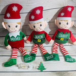 Può includere: Tre bambole elfo di peluche con cappelli rossi con fiocchi di neve bianchi, abiti verdi e rossi e gambe a righe. Ogni bambola ha un nome ricamato sul petto: "MIMI", "JAXON" e "DANNY". L'anno "2022" è su ogni piede. È presente anche un piccolo albero di Natale verde glitterato.