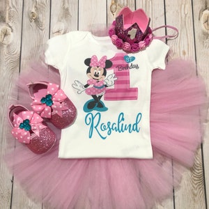 Może przedstawiać: R&oacute;żowa sp&oacute;dniczka tiulowa, biały body z numerem jeden w r&oacute;żowo-białe paski, grafiką Minnie Mouse oraz tekstem "1. urodziny" i "Rosalind". Body ma r&oacute;wnież r&oacute;żową kokardkę na kołnierzyku. Str&oacute;j uzupełnia r&oacute;żowa korona z srebrną jedynką i r&oacute;żowymi kwiatami.