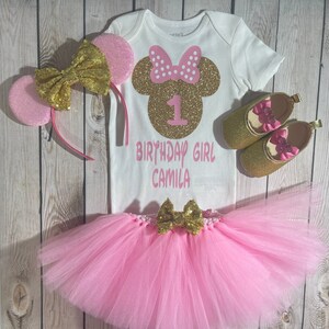 May include: A pink and gold Minnie Mouse themed first birthday outfit for a baby girl. The outfit includes a white bodysuit with a gold glitter Minnie Mouse head and the text "1 Birthday Girl Camila", a pink tulle tutu with a gold bow, a pink headband with gold glitter ears, and a pair of gold glitter shoes with pink bows.