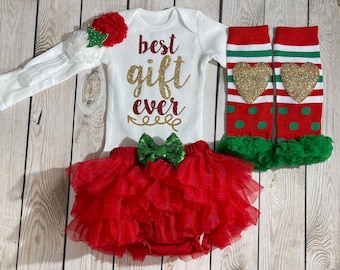 Puede incluir: Un conjunto de ropa para bebé que incluye un body blanco con el texto "Best Gift Ever" en purpurina roja y dorada, un tutú rojo, un lazo verde con lentejuelas, calentadores de piernas con corazones dorados con purpurina y una diadema a juego.