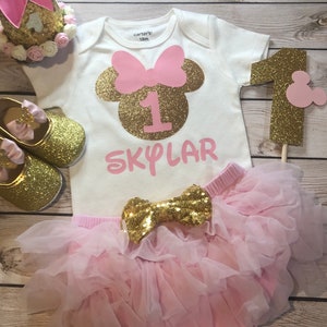 Puede incluir: Un conjunto de primer cumplea&ntilde;os de Minnie Mouse en rosa y dorado para una ni&ntilde;a. El conjunto incluye un body blanco con una cabeza de Minnie Mouse de purpurina dorada, un lazo rosa y el n&uacute;mero uno. El body tambi&eacute;n tiene el nombre "SKYLAR" escrito en rosa. El conjunto tambi&eacute;n incluye una falda de tul rosa con un lazo de purpurina dorada.