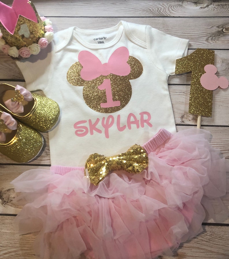 Puede incluir: Un conjunto de primer cumplea&ntilde;os de Minnie Mouse en rosa y dorado para una ni&ntilde;a. El conjunto incluye un body blanco con una cabeza de Minnie Mouse de purpurina dorada, un lazo rosa y el n&uacute;mero uno. El body tambi&eacute;n tiene el nombre "SKYLAR" escrito en rosa. El conjunto tambi&eacute;n incluye una falda de tul rosa con un lazo de purpurina dorada.