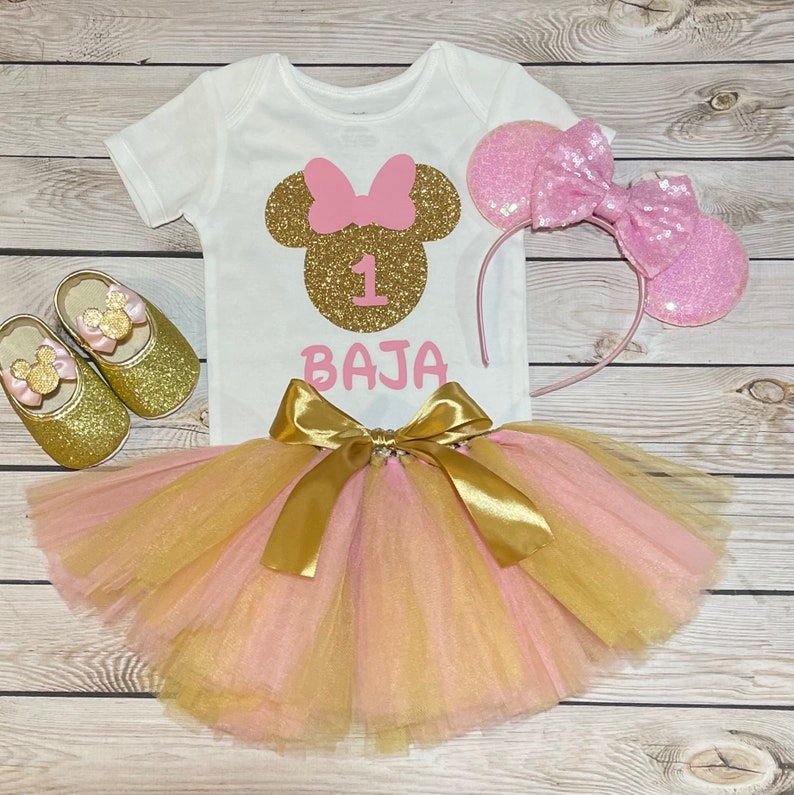 May include: A pink and gold Minnie Mouse themed first birthday outfit for a baby girl. The outfit includes a white t-shirt with a gold glitter Minnie Mouse head, a pink bow, and the number "1" in gold glitter. The outfit also includes a pink and gold tulle tutu and a pink headband with Minnie Mouse ears. The t-shirt also has the name "BAJA" written in pink.