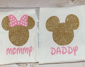 May include: Two white t-shirts with gold glitter Mickey Mouse silhouettes. The left shirt has a pink bow on top of the head and the word "mommy" in pink letters. The right shirt has the word "daddy" in pink letters.