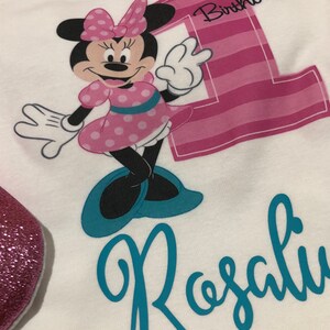 Może przedstawiać: Biały śpioszek z numerem jeden w paski r&oacute;żowo-białe, Minnie Mouse w r&oacute;żowo-białe kropki i tekstem "1. urodziny". Imię "Rosalie" jest napisane turkusowym kursywą pod Minnie Mouse.