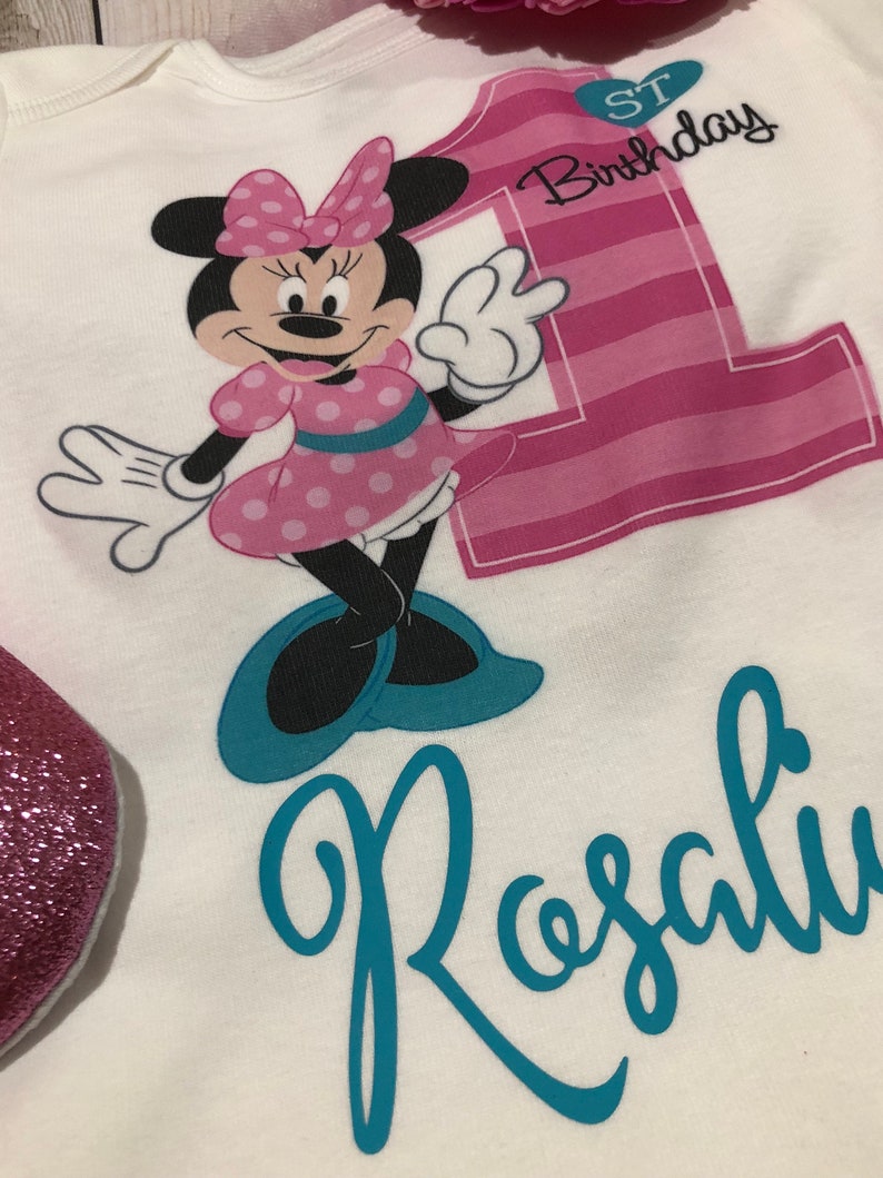 Może przedstawiać: Biały śpioszek z numerem jeden w paski r&oacute;żowo-białe, Minnie Mouse w r&oacute;żowo-białe kropki i tekstem "1. urodziny". Imię "Rosalie" jest napisane turkusowym kursywą pod Minnie Mouse.