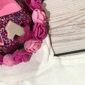 May include: A pink and white glittery crown with a white number one on top. The crown is decorated with pink felt flowers.