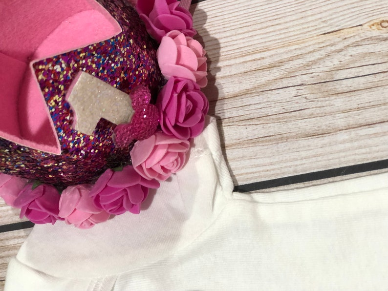 May include: A pink and white glittery crown with a white number one on top. The crown is decorated with pink felt flowers.