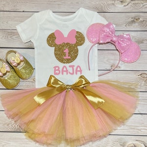 May include: A pink and gold Minnie Mouse themed first birthday outfit for a baby girl. The outfit includes a white t-shirt with a gold glitter Minnie Mouse head, a pink bow, and the number 1. The t-shirt also has the name "BAJA" written in pink glitter. The outfit also includes a pink and gold tulle tutu skirt, a pink headband with glitter Minnie Mouse ears, and a pair of gold glitter shoes with Minnie Mouse ears.