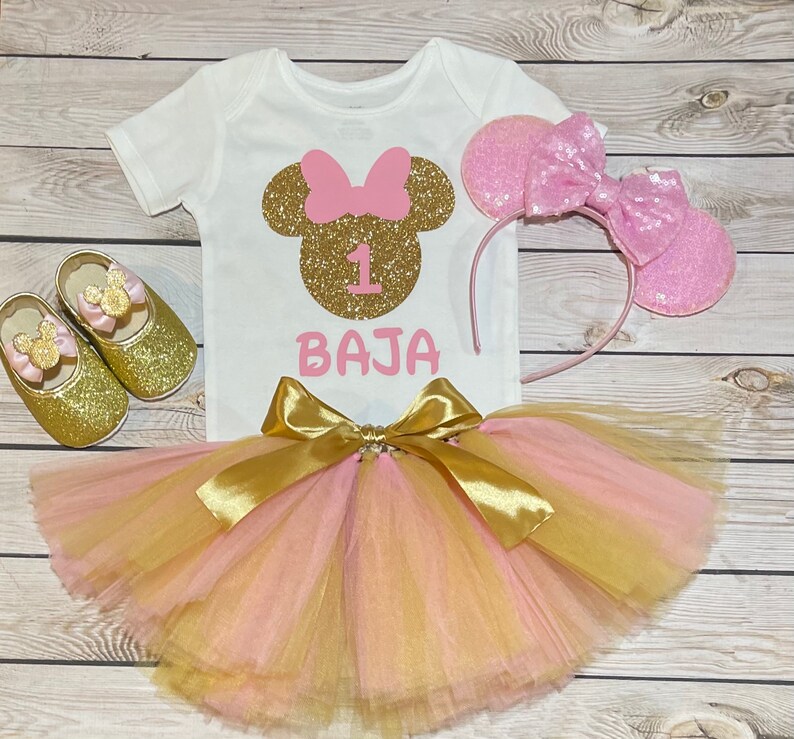 May include: A pink and gold Minnie Mouse themed first birthday outfit for a baby girl. The outfit includes a white t-shirt with a gold glitter Minnie Mouse head, a pink bow, and the number 1. The t-shirt also has the name "BAJA" written in pink glitter. The outfit also includes a pink and gold tulle tutu skirt, a pink headband with glitter Minnie Mouse ears, and a pair of gold glitter shoes with Minnie Mouse ears.