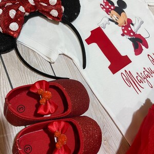 May include: A red and white Minnie Mouse themed outfit for a baby girl. The outfit includes a white onesie with a red number one and Minnie Mouse graphic, a red tulle skirt, a red glitter headband with a red and white polka dot bow, and red glitter shoes with a Minnie Mouse bow.